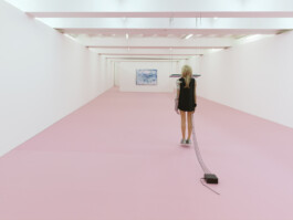 Eine Puppe hängt mit ihrem Kinn an einer dicken Glasscheibe. Ihre Füße schweben kurz über dem Boden. Der Raum ist ein weißer langer Schlauch mit rosa Boden.