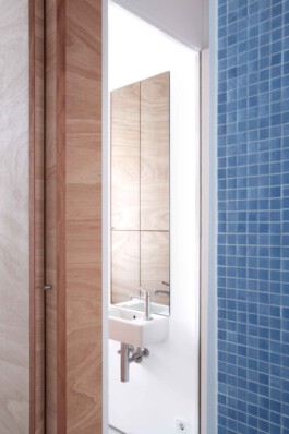 Kleines Waschbecken mit Spiegel, in dem man Fronten aus Tischlerplatte sieht. Im Vordergrund blaue Fliesen. 