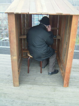 Mann sitzt in einer einfachen Hütte und beobachtet durch ein Fenster die Stadt 