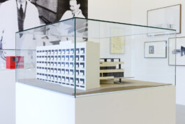 Architekturmodell unter einer Glashaube, im Hintergrund Zeichnungen und Fotografien 