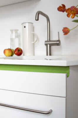 Detailaufnahme einer Küche mit einer weißen Arbeitsplatte aus Corian, weißen Fronten und einer grünen Griffleiste. 