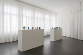 Heller Raum mit dunklem Boden, umlaufend weißen Vorhängen und großen weißen Sockeln auf denen kleine bunte Glasobjekte stehen 