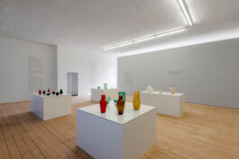 Großer Raum mit Holzdielen, hellgrauen Stellwänden und großen weißen Sockeln auf denen kleine bunte 