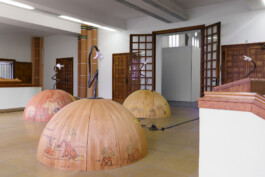 Drei Halbkugeln im Vorraum einer Kirche. Am höchsten Punkt ist auf jeder Kugel jeweils eine organisch verbogene Stahlstange befestigt, an deren Spitze eine Hand mit ausgestrecktem Zeigefinger in einem weißen Handschuh sitzt.