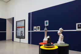 Drei Objekte auf runten hohen Sockeln im Vordergrund, dahinter Bilder auf dunkelblauen und weißen Flächen an der Wand 
