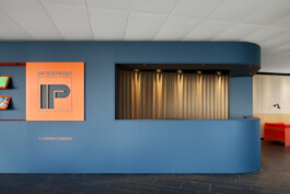 Foyer-Ausbau mit rundem Tresen und Backoffice für die Interprint Group in dunkelblau. Einsatz aus orange pulverbeschichteten 3mm Stahl mit Firmenlogo. 