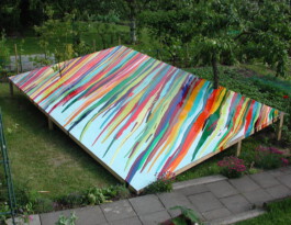 Installation aus einer rechteckigen schrägen Fläche mit Streifen aus verlaufenen bunten Farben in einem Garten. Die Bäume scheinen durch die Fläche zu wachsen. 