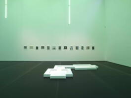 Weißes flaches Objekt auf dunklem Boden in einem sehr hohen Raum. Im Hintergrund kleine Bilder an der Wand. 