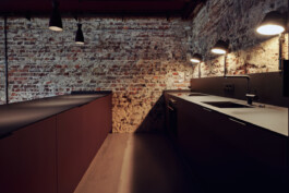 Zweizeilige Küche mit dunkler super-matter Oberfläche aus Linoleum und HPL vor unverputztem Mauerwerk.