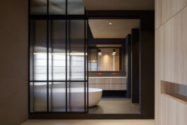 Blick in dunkles Badezimmer mit Waschtisch aus Weißtanne, freistehender Badewanne und Schiebetür aus Strukturglas. 