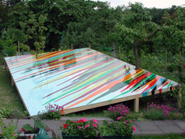 Installation aus einer rechteckigen schrägen Fläche mit Streifen aus verlaufenen bunten Farben in einem Garten. Die Bäume scheinen durch die Fläche zu wachsen.